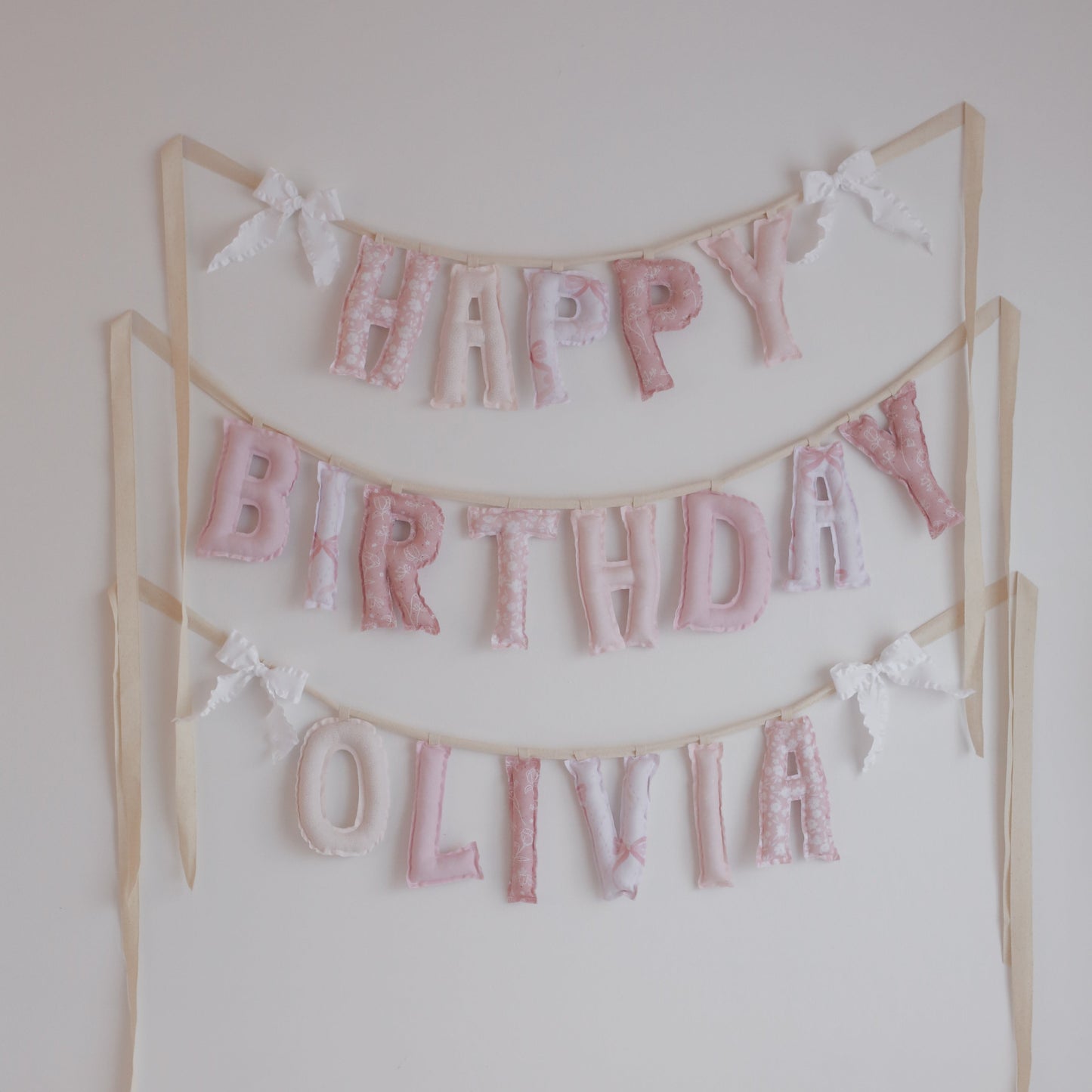 Lace & Petals handmade ‘Happy Birthday’ garland by Social Heirloom – soft pinks, dusty rose, and bow-print fabrics with white ruffle bows, perfect for first birthdays, nurseries, or delicate, heirloom celebrations. Shown here with name addition Olivia.