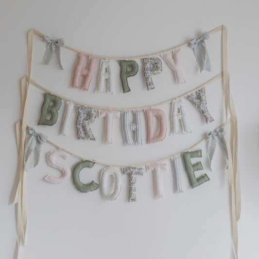 Blush & Olive handmade ‘Happy Birthday’ garland by Social Heirloom – soft pinks, sage greens, florals, and neutrals with lavender ruffle bows, perfect for first birthdays, nurseries, and timeless heirloom celebrations. Shown with name addition Scottie.