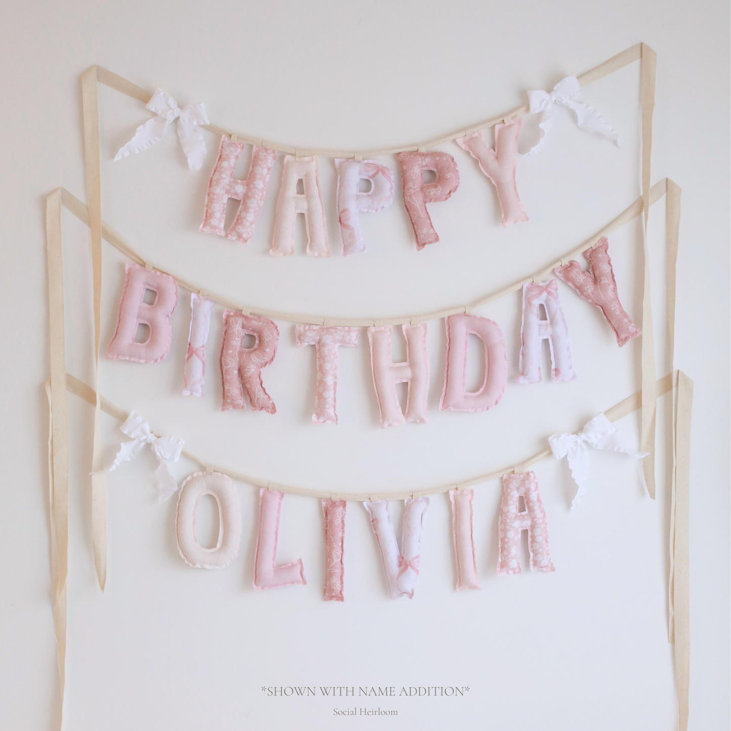 Lace & Petals birthday garland by Social Heirloom – handmade pink fabric letters in bow and floral prints with white ruffle bows, designed for a soft, storybook-style birthday or nursery display. Shown with name addition Olivia.
