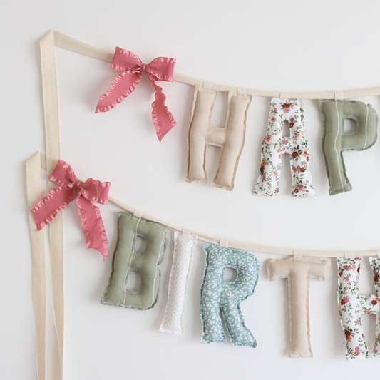 Close-up of Whispering Blooms birthday garland by Social Heirloom – hand-sewn cotton letters in soft greens, florals, and neutrals with raspberry pink ruffle bows, ideal for heirloom celebrations and delicate nursery styling.