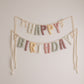 A pastel 'HAPPY BIRTHDAY' fabric banner with textured letters hanging from two strings against a plain off-white wall.