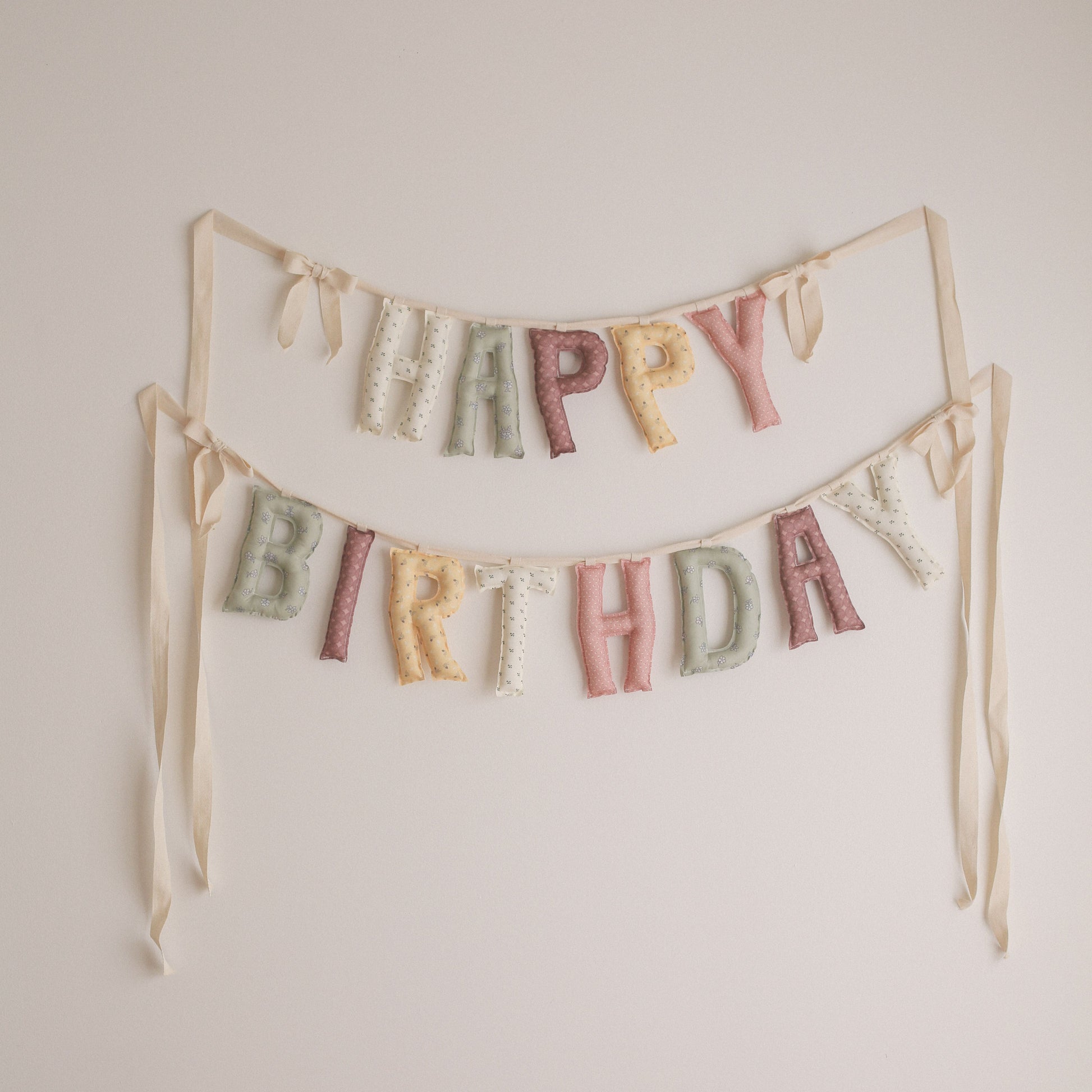 A pastel 'HAPPY BIRTHDAY' fabric banner with textured letters hanging from two strings against a plain off-white wall.