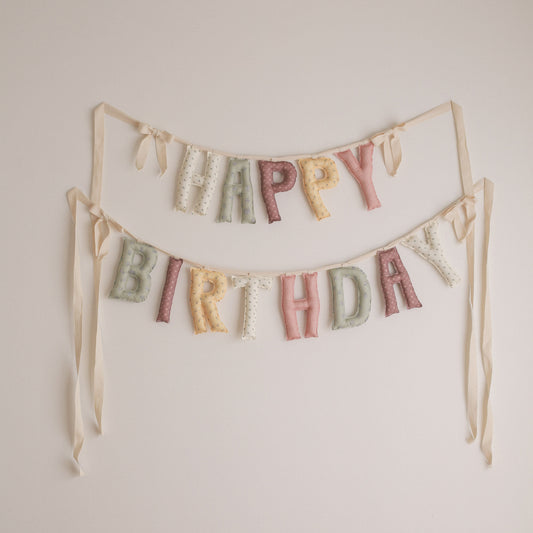 A pastel 'HAPPY BIRTHDAY' fabric banner with textured letters hanging from two strings against a plain off-white wall.