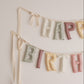 A pastel fabric letter banner spelling HAPPY BIRTH hanging on a light neutral wall, with cream ribbons and soft multicolor letters including pale green, dusty pink, cream, and mustard.