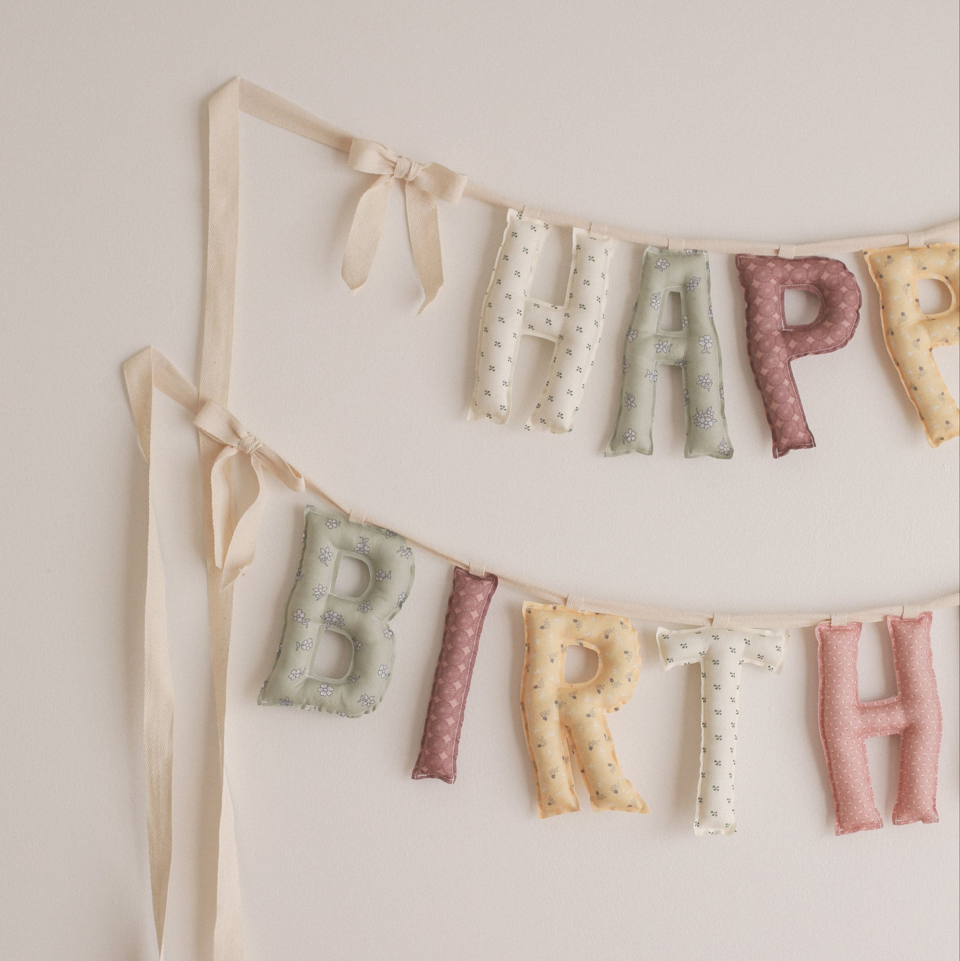 A pastel fabric letter banner spelling HAPPY BIRTH hanging on a light neutral wall, with cream ribbons and soft multicolor letters including pale green, dusty pink, cream, and mustard.