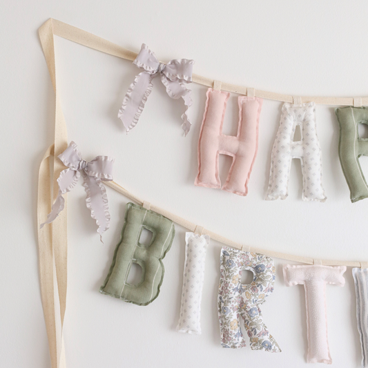 Close-up of Blush & Olive birthday garland by Social Heirloom – hand-sewn cotton letters in soft floral, blush, olive, and neutral prints with delicate grey ruffle bows, ideal for nursery decor or vintage-inspired birthdays.