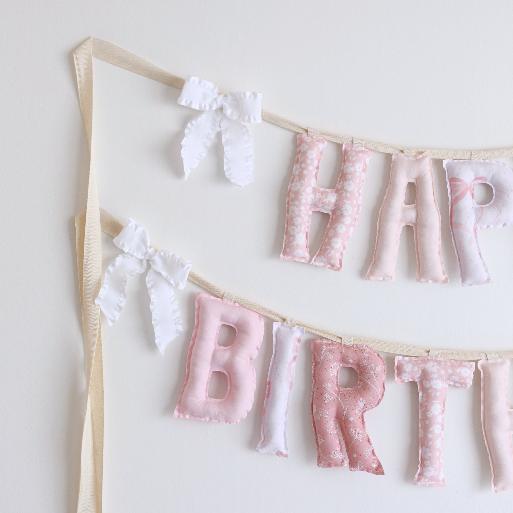 Close-up of Lace & Petals birthday garland by Social Heirloom – handmade pink fabric letters in bow and floral prints with white ruffle bows, designed for a soft, storybook-style birthday or nursery display