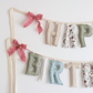 Close-up of Whispering Blooms birthday garland by Social Heirloom – hand-sewn cotton letters in soft greens, florals, and neutrals with raspberry pink ruffle bows, ideal for heirloom celebrations and delicate nursery styling.