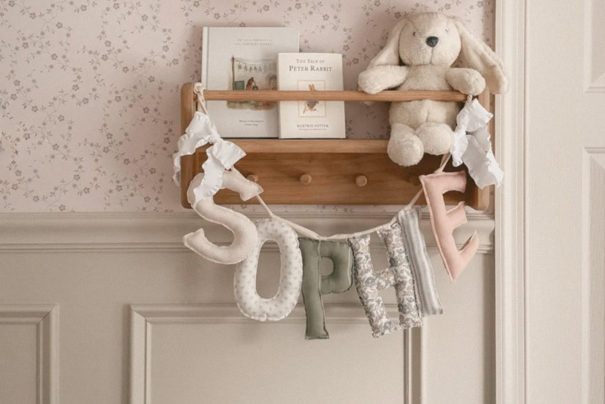 Wooden shelf with decorative items including a personalized fabric name garland, a plush toy, and books in a nursery setting.