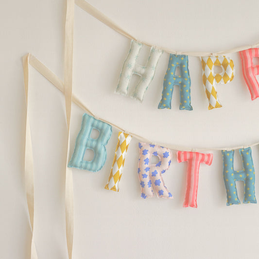 Close up of the Wind-Up Circus Happy Birthday garland, soft filled fabric letters in stars, harlequin diamonds, coral stripes and little blue flowers